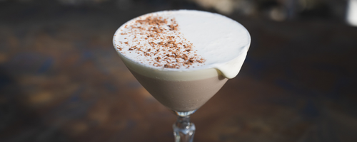 A creamy Mocha Martini topped with coffee foam and a dusting of cinnamon, made with Damn Fine Vodka and Mocha Bourbon Cream Liqueur, served in a chilled coupe glass surrounded by warm holiday light.